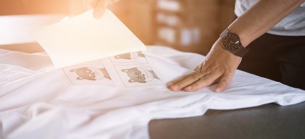 Young woman pull out paper from waterproof film on fabric. worker working on manual screen printing on t-shirt at her shop.